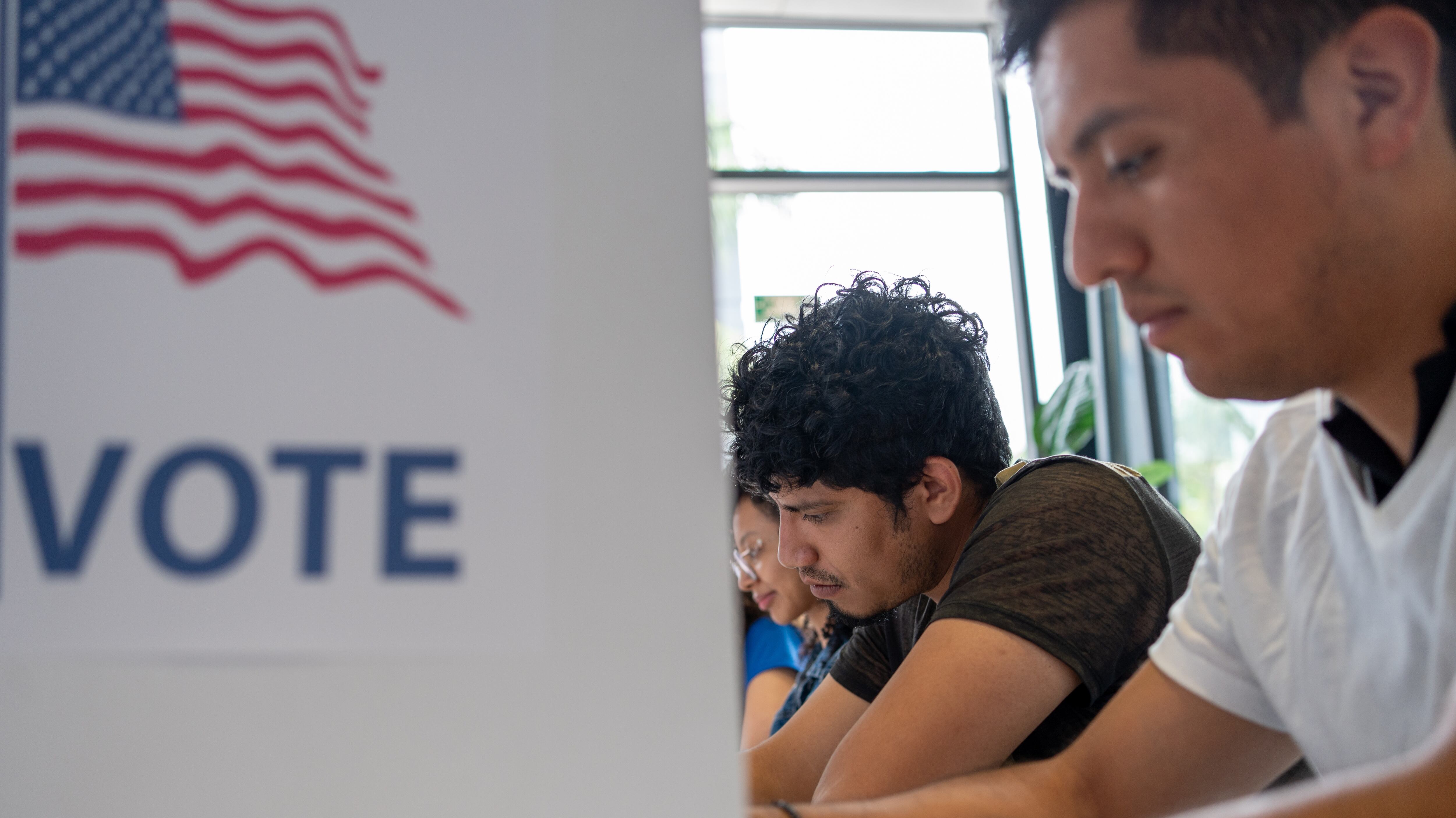 Hispanic young man is seen intently casting his vote in a U.S. election. The voting booth is adorned with an American flag and the word "VOTE” democracy, voting rights, and electoral participation.